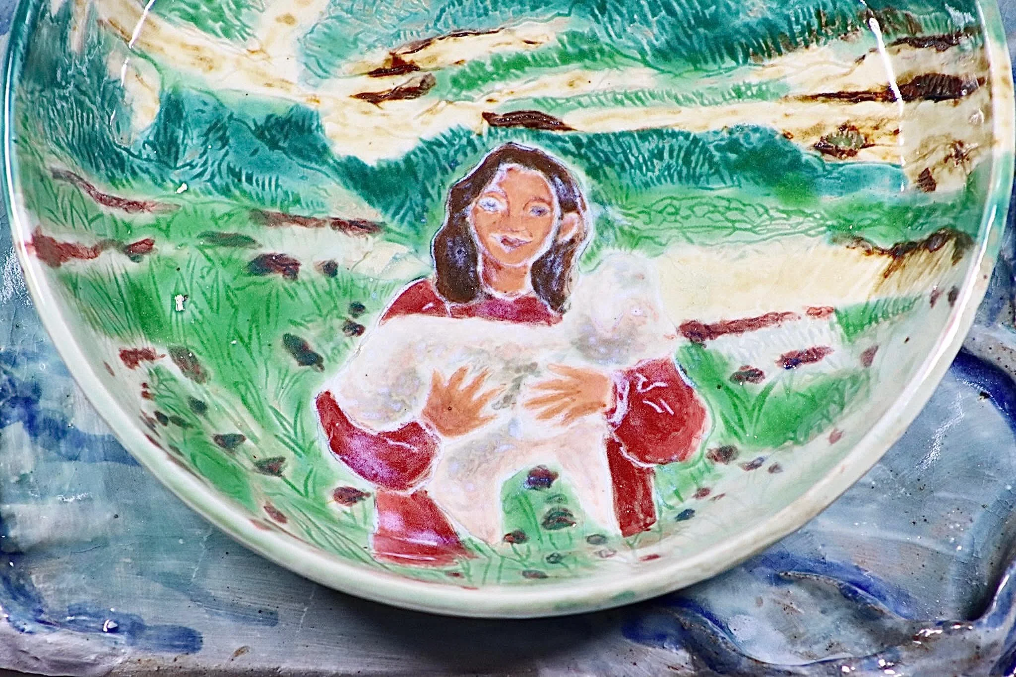 "My Rushing Waves of Culture II", 2026. Glazed Red Stoneware and Porcelain. 10.75" x 8.25" inches.

 Part of a series . A ceramic plate with a tray in the form of a lake showcasing my mother in San Jose, Peru cradling a sheep.