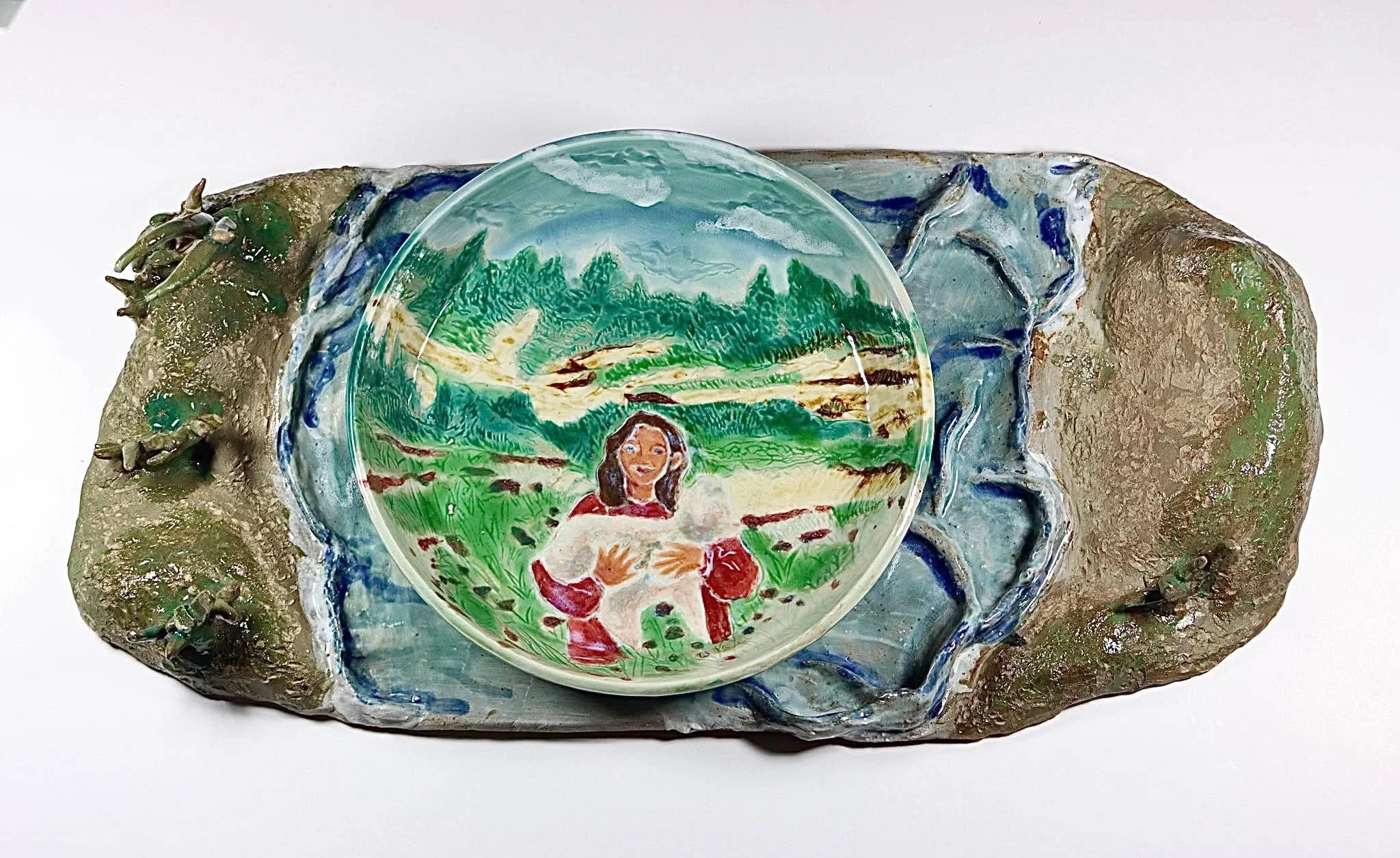 "My Rushing Waves of Culture II", 2026. Glazed Red Stoneware and Porcelain. 10.75" x 8.25" inches.

 Part of a series . A ceramic plate with a tray in the form of a lake showcasing my mother in San Jose, Peru cradling a sheep.