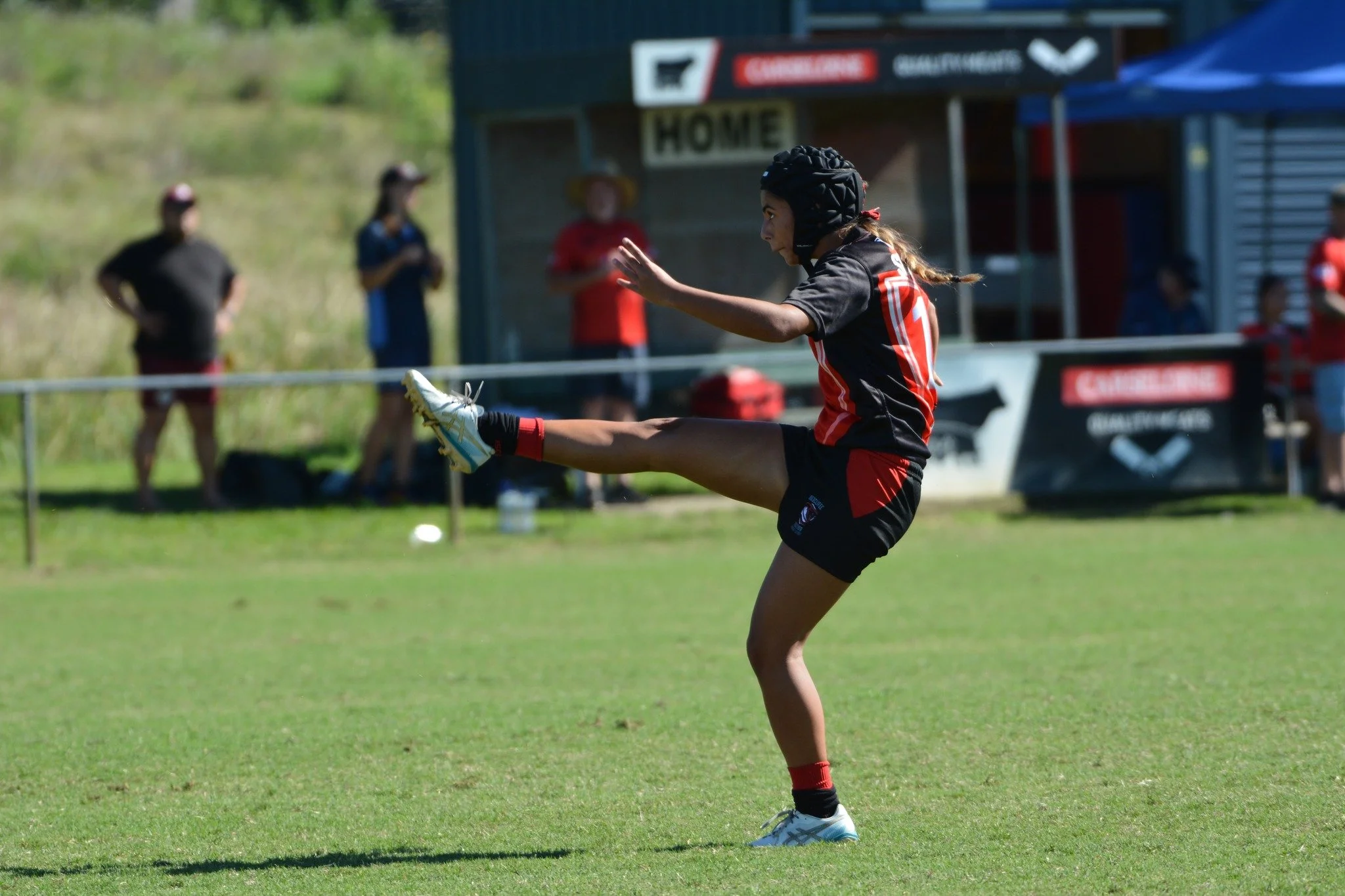A young girl wearing a black and red sports uniform, helmet, and cleats is playing rugby, kicking the ball on a grassy field during a match or practice, with spectators and a building in the background.