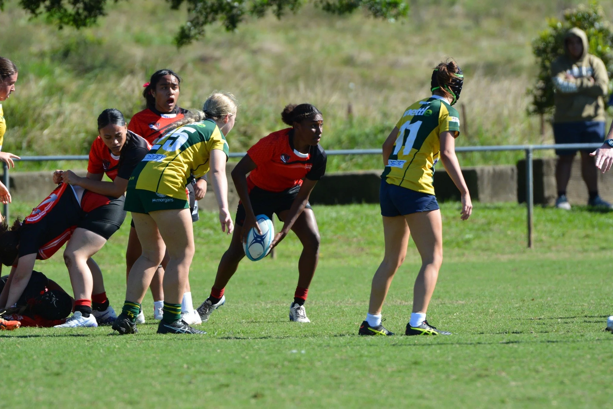 Women playing rugby on a grassy field, with some players in red and others in yellow and blue jerseys, preparing for a play.