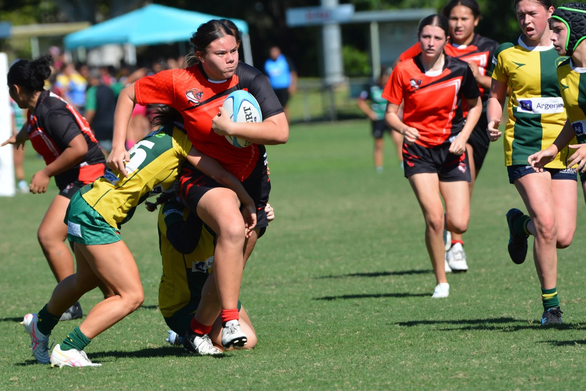 Girls playing rugby, one girl in a red and black jersey carrying a rugby ball while being tackled by a girl in a yellow and green jersey on a grassy field.