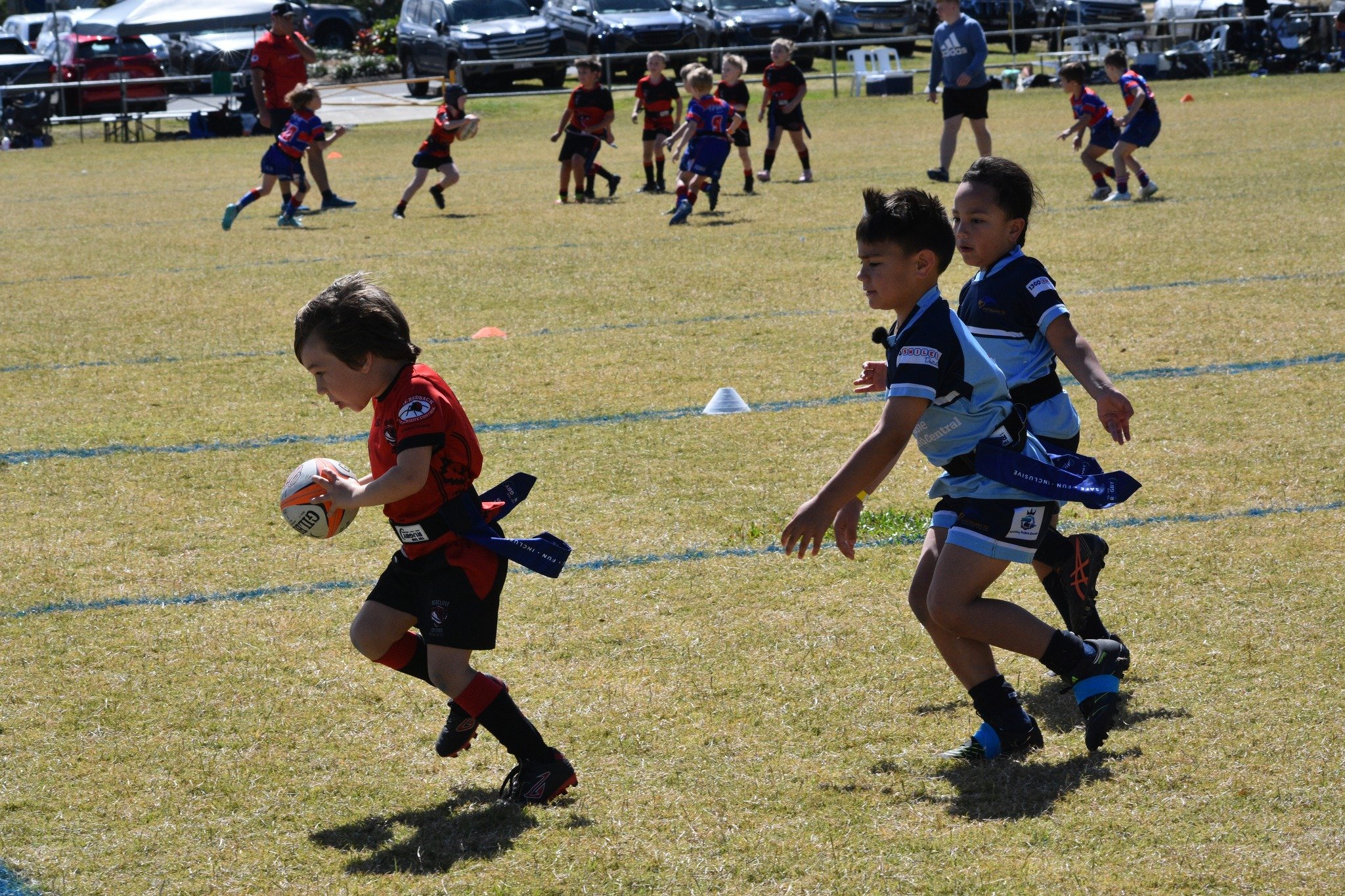 Young boys playing rugby on a grassy field during a youth sports game, with others in the background, some running and some standing, in sunny weather.