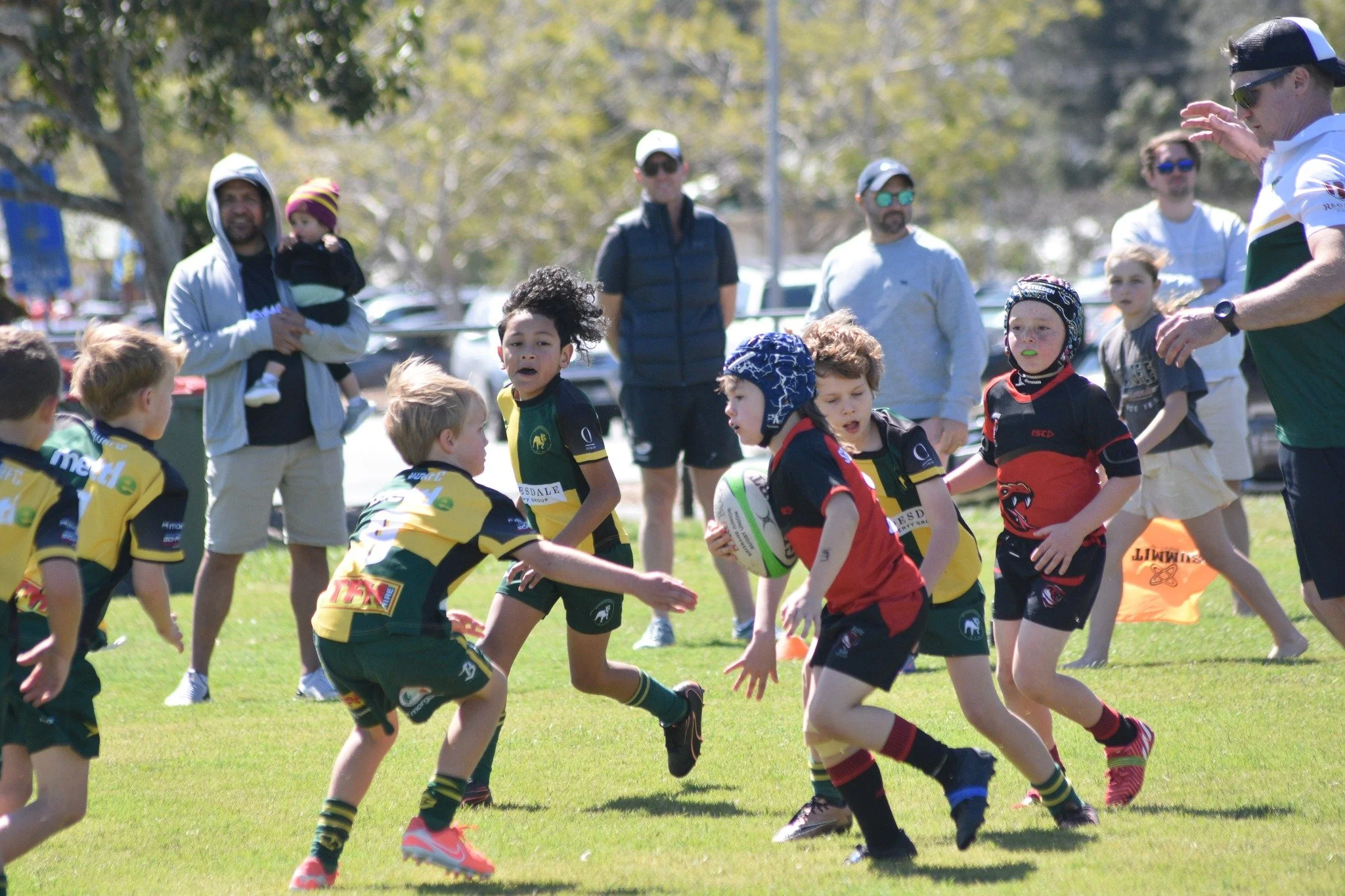 Young children playing rugby on a grassy field, wearing colorful uniforms and protective headgear, with adults watching in the background.