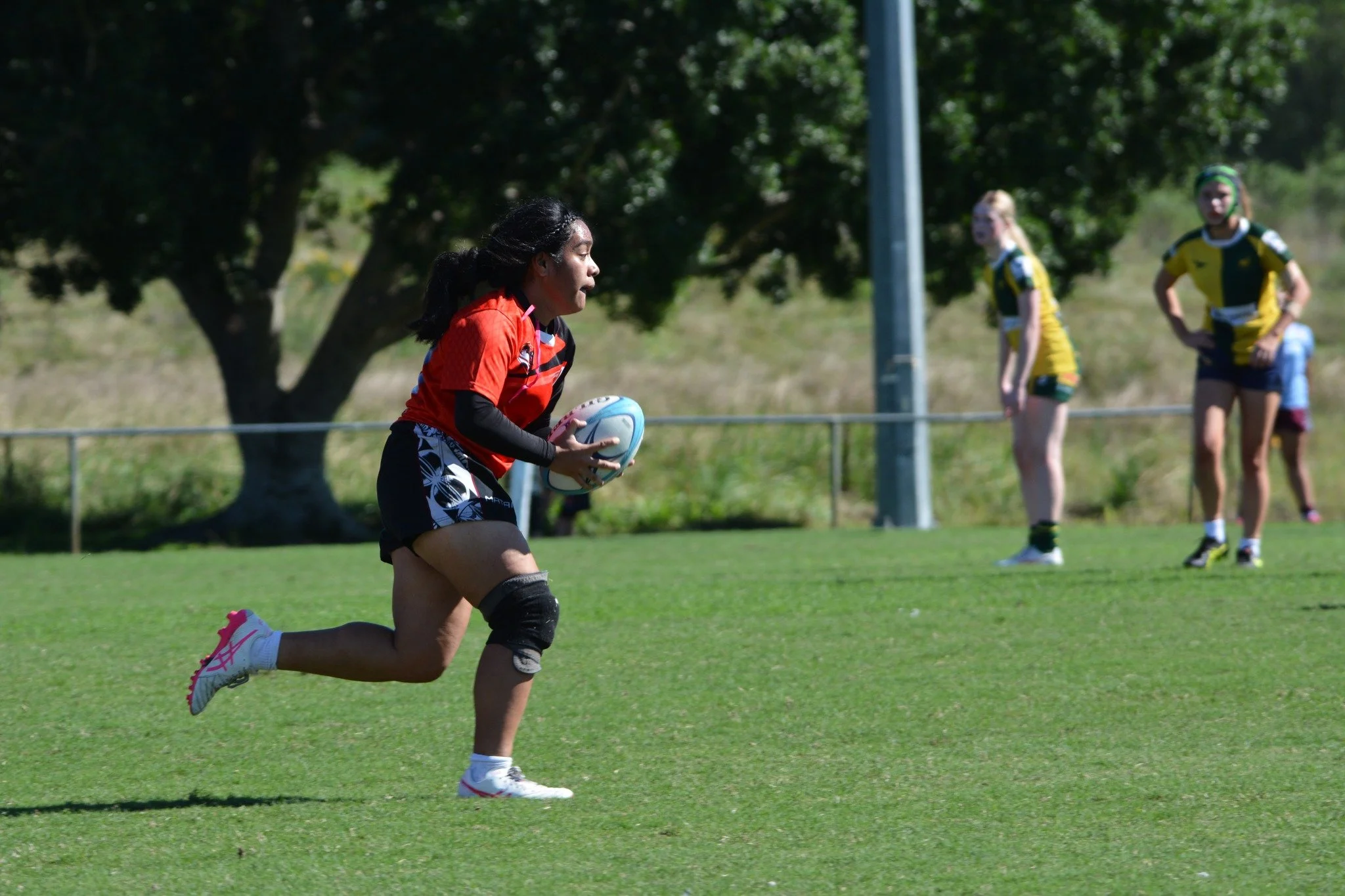 A woman in a red sports jersey and black shorts running on a rugby field while carrying a rugby ball, with three women in athletic gear in the background
