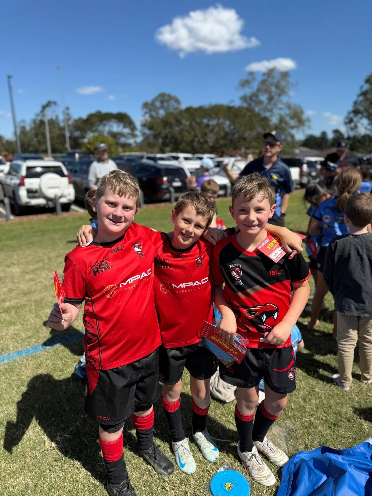Three young boys in red and black sports uniforms standing together outdoors, smiling, with other children and adults in the background during a sunny day at a sports event.