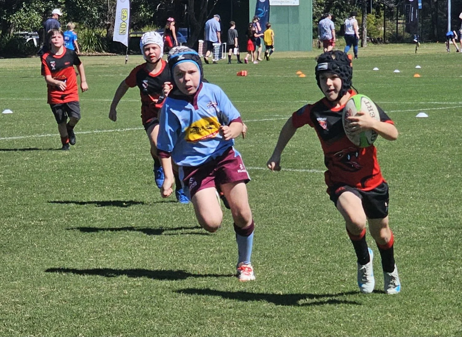 Young children playing rugby on a grassy field, running with a ball, with others in the background during a sports event.