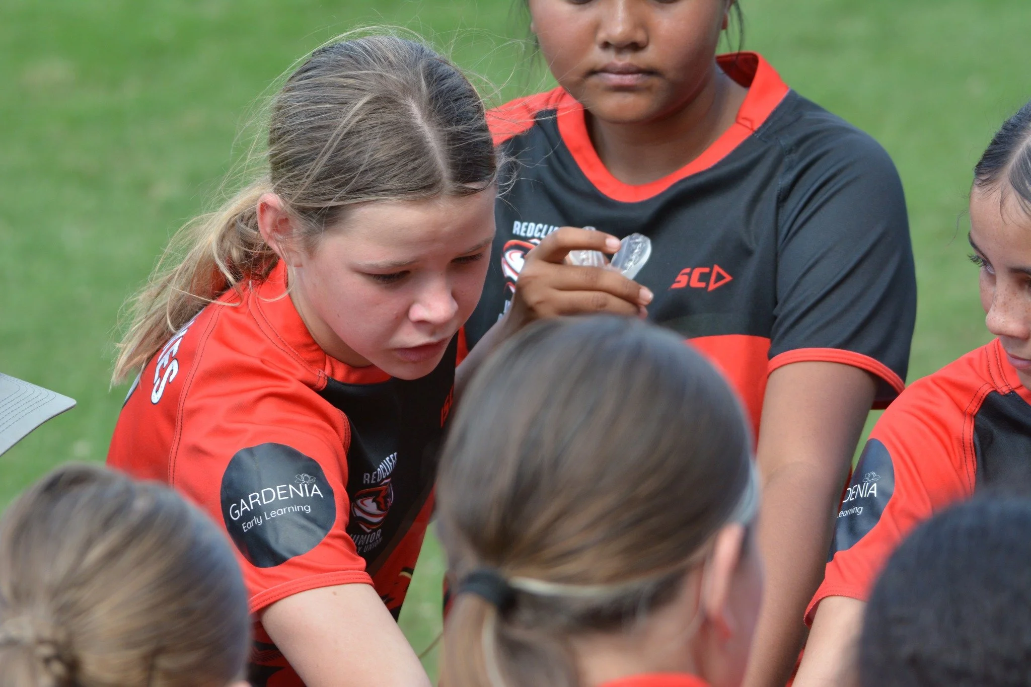 Group of young female athletes in red and black sports uniforms gathered in a huddle, outdoors on a grass field during a team meeting or strategy session.