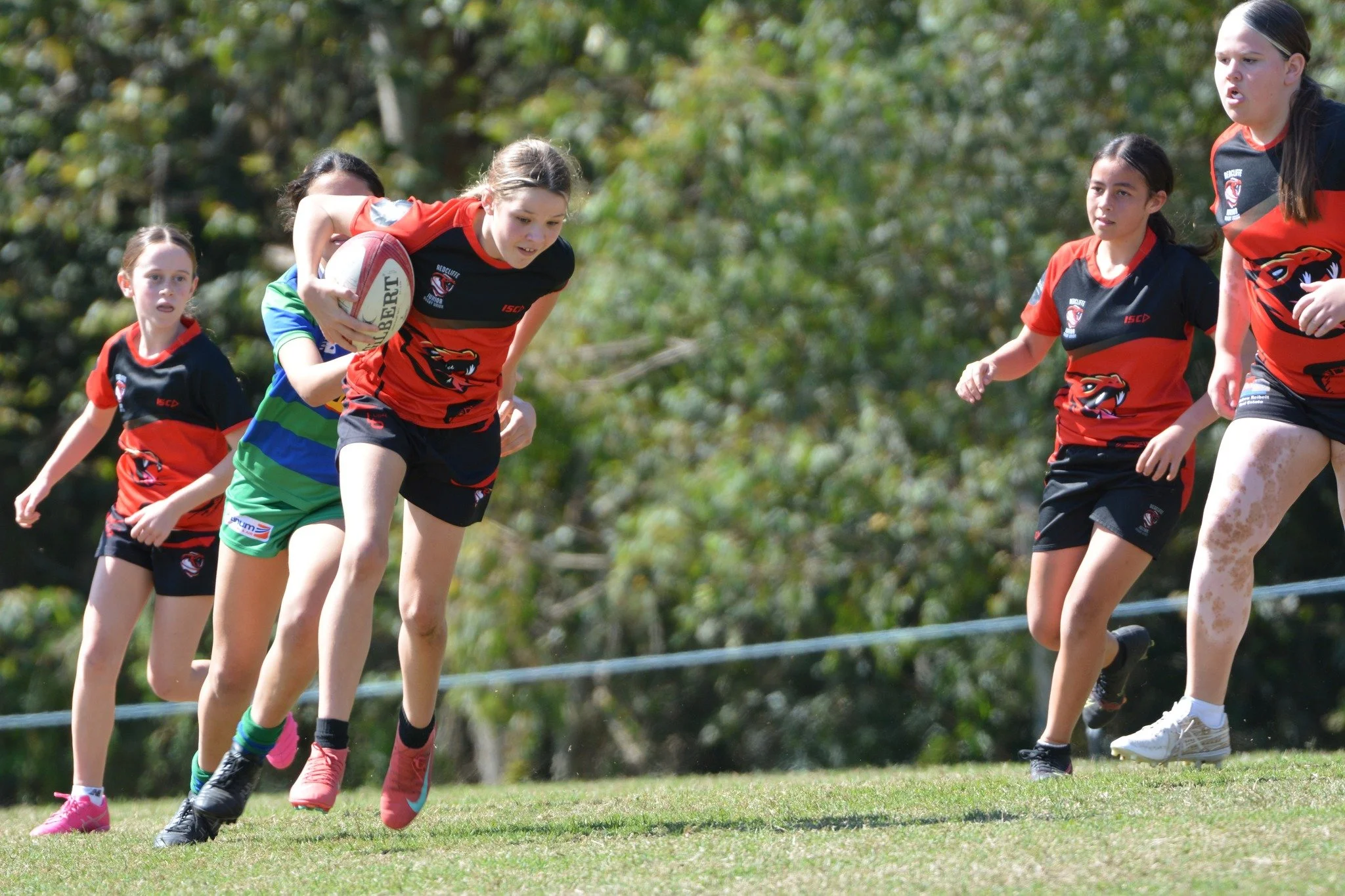 Young girls playing rugby, with one girl running with a rugby ball as others chase her on a grassy field during daytime.