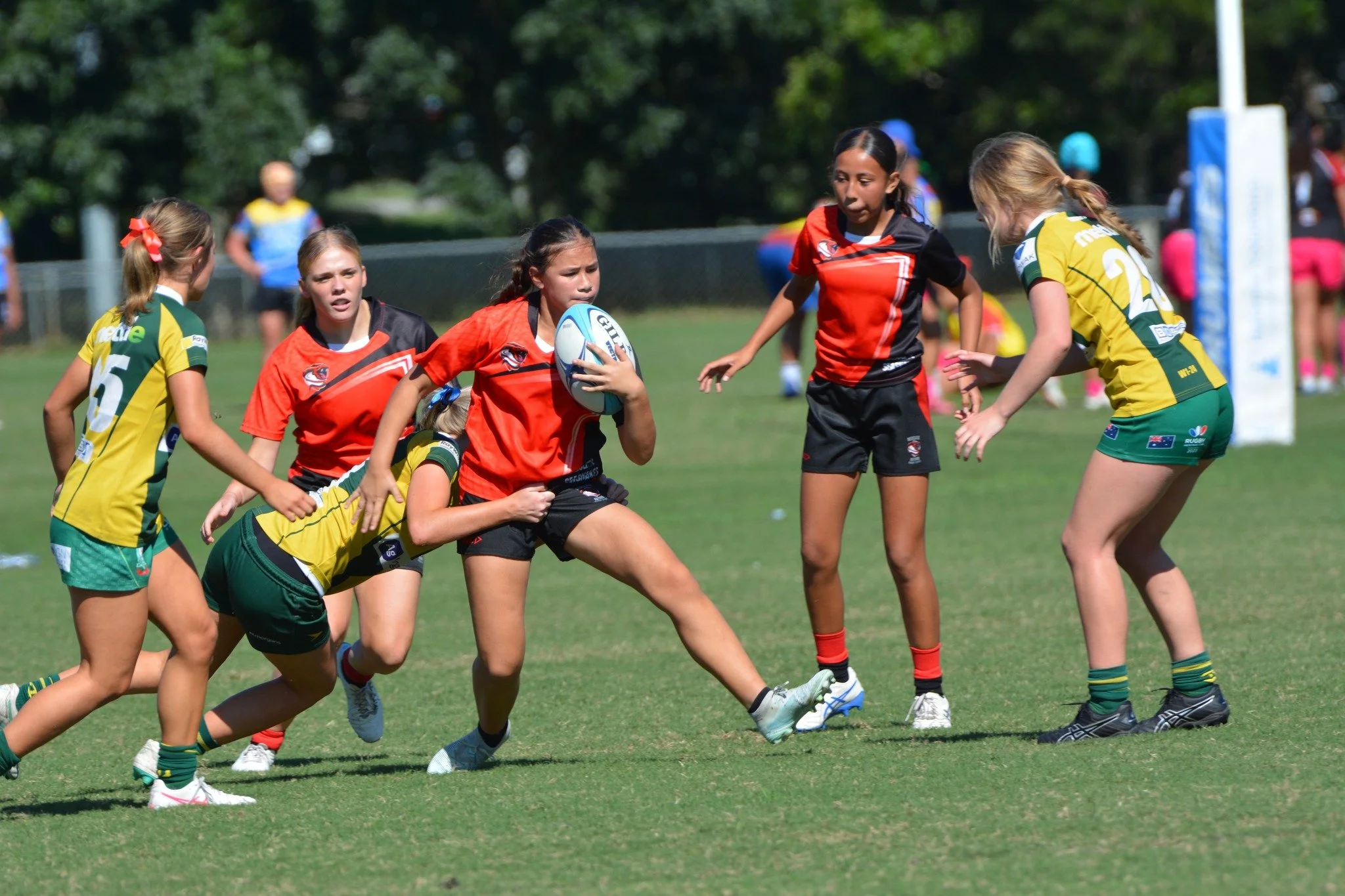 Girls playing rugby on a grassy field, with one girl holding a rugby ball and others in position to make a play, in bright sports uniforms.