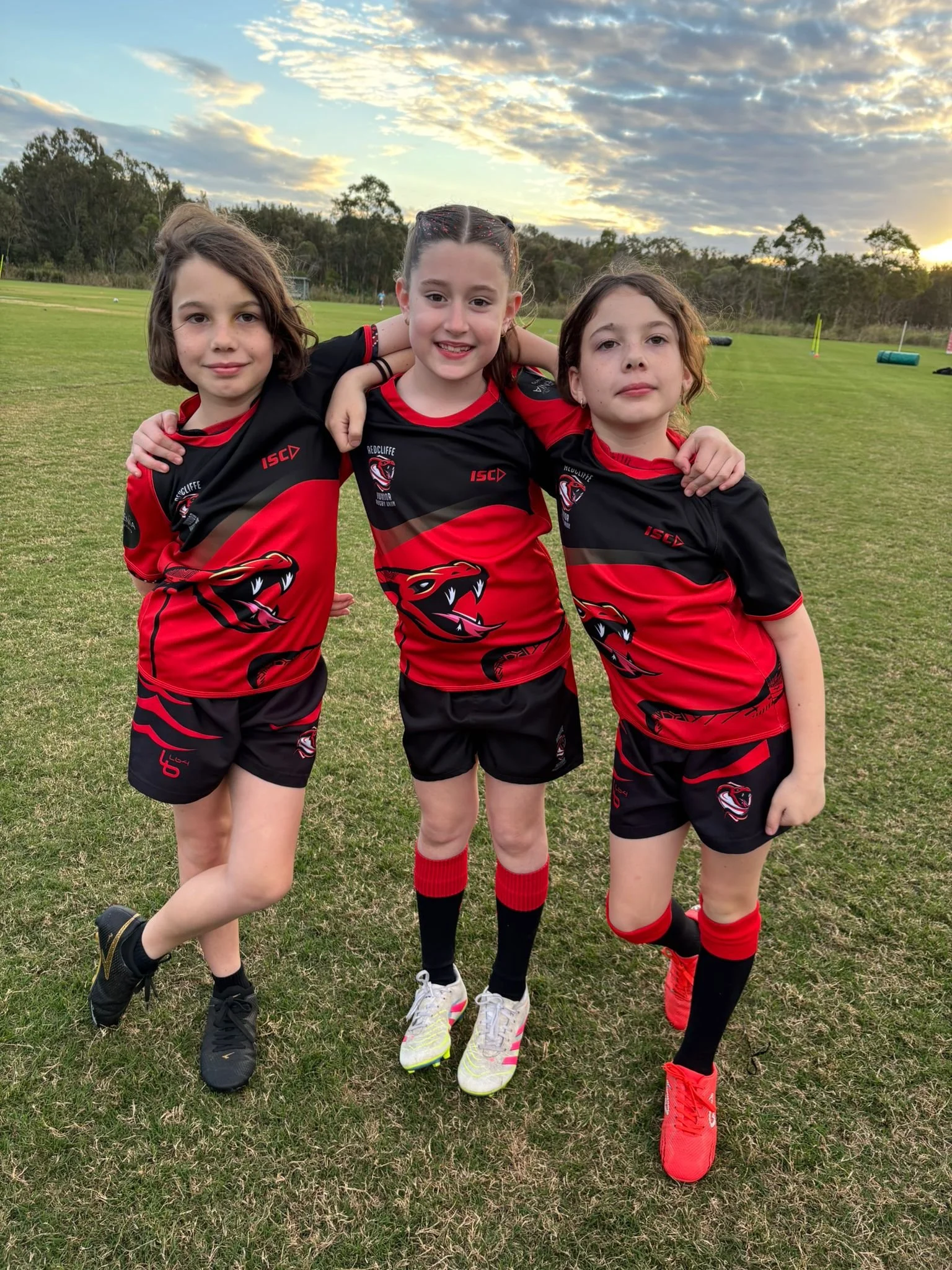 Three young girls in red and black sports uniforms stand on a field with their arms around each other, smiling at the camera.