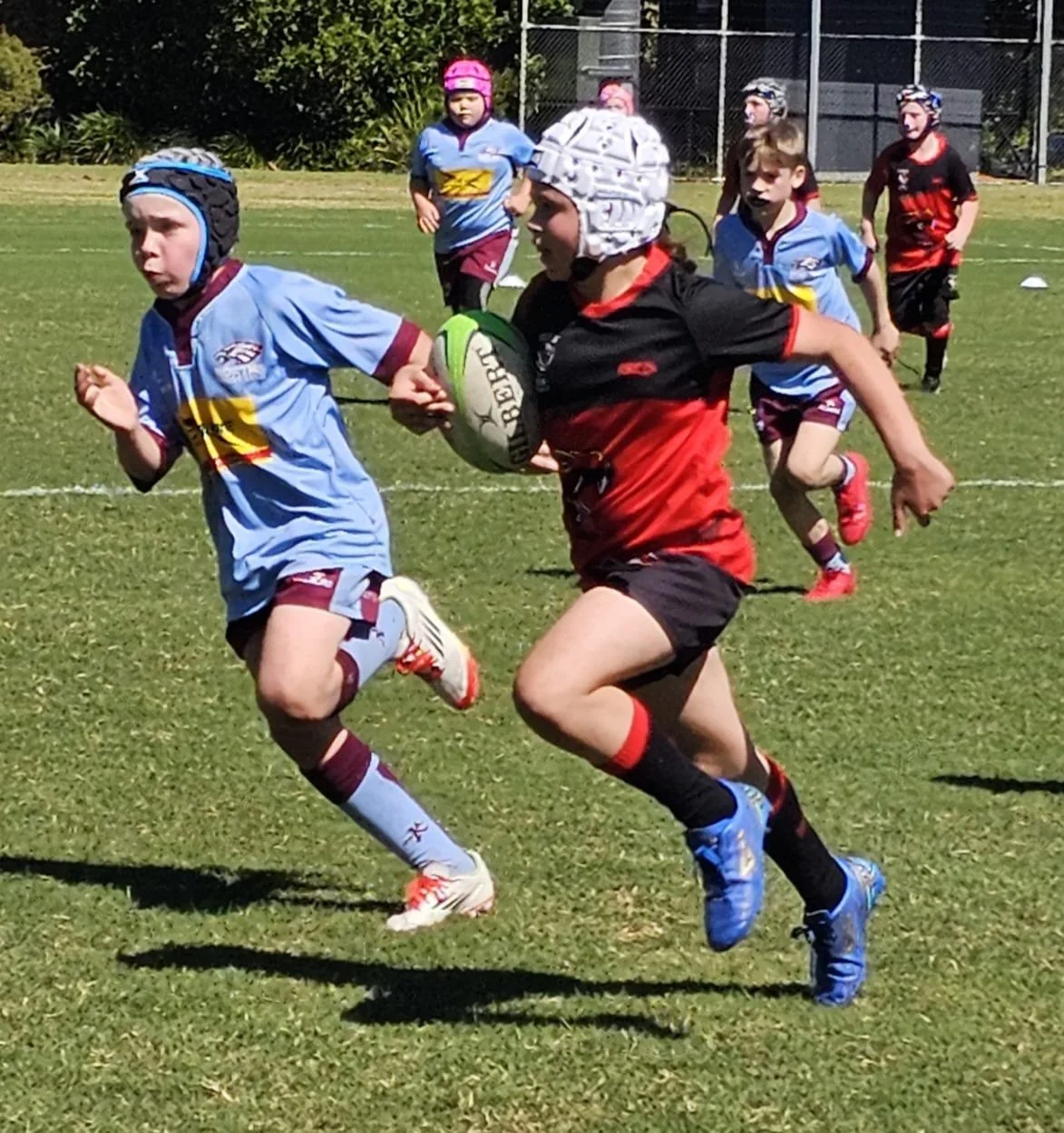 Children playing rugby on a grassy field, some wearing protective headgear, with a focus on a girl in a black and red uniform running with the ball surrounded by other kids in blue and red uniforms.
