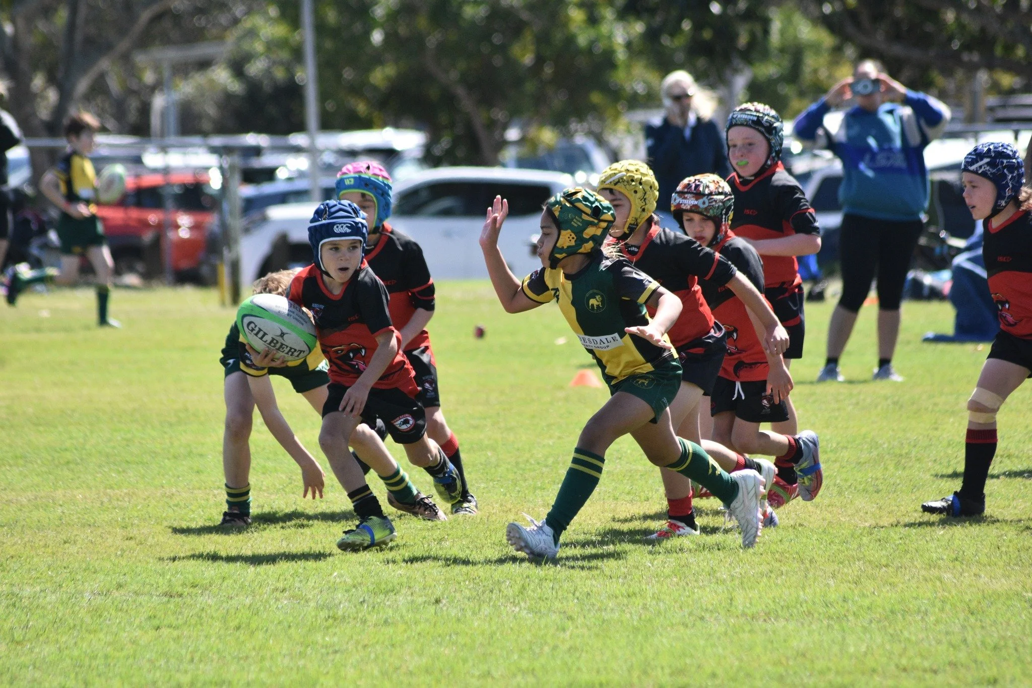 Young children playing rugby on a grassy field, some wearing helmets and sports uniforms, with a few adults and parked cars in the background.