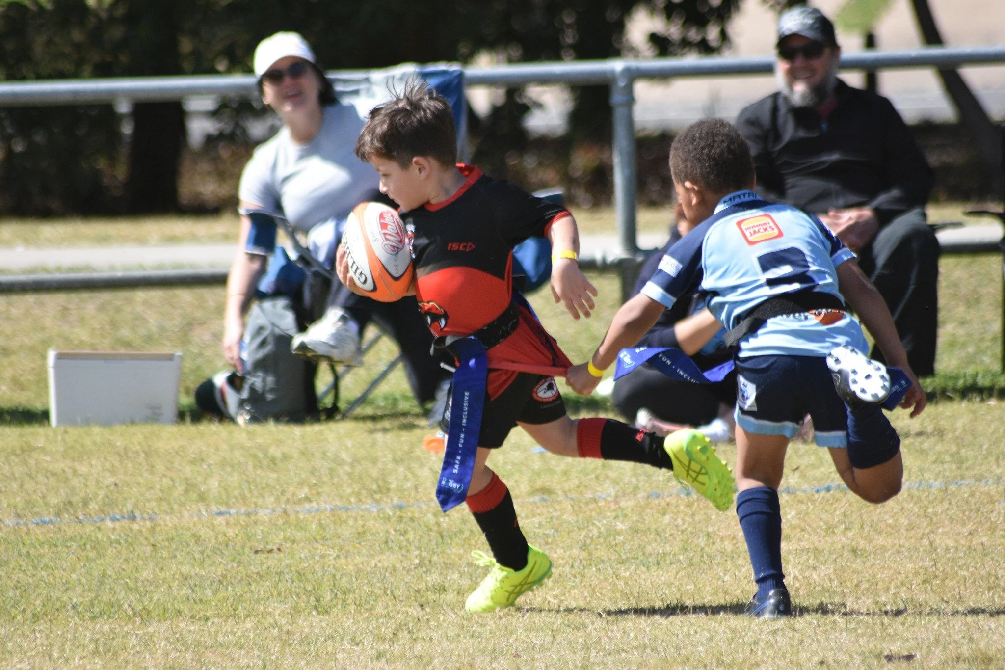 Two young boys playing rugby on a grassy field, with two adults sitting on the sidelines watching in the background.
