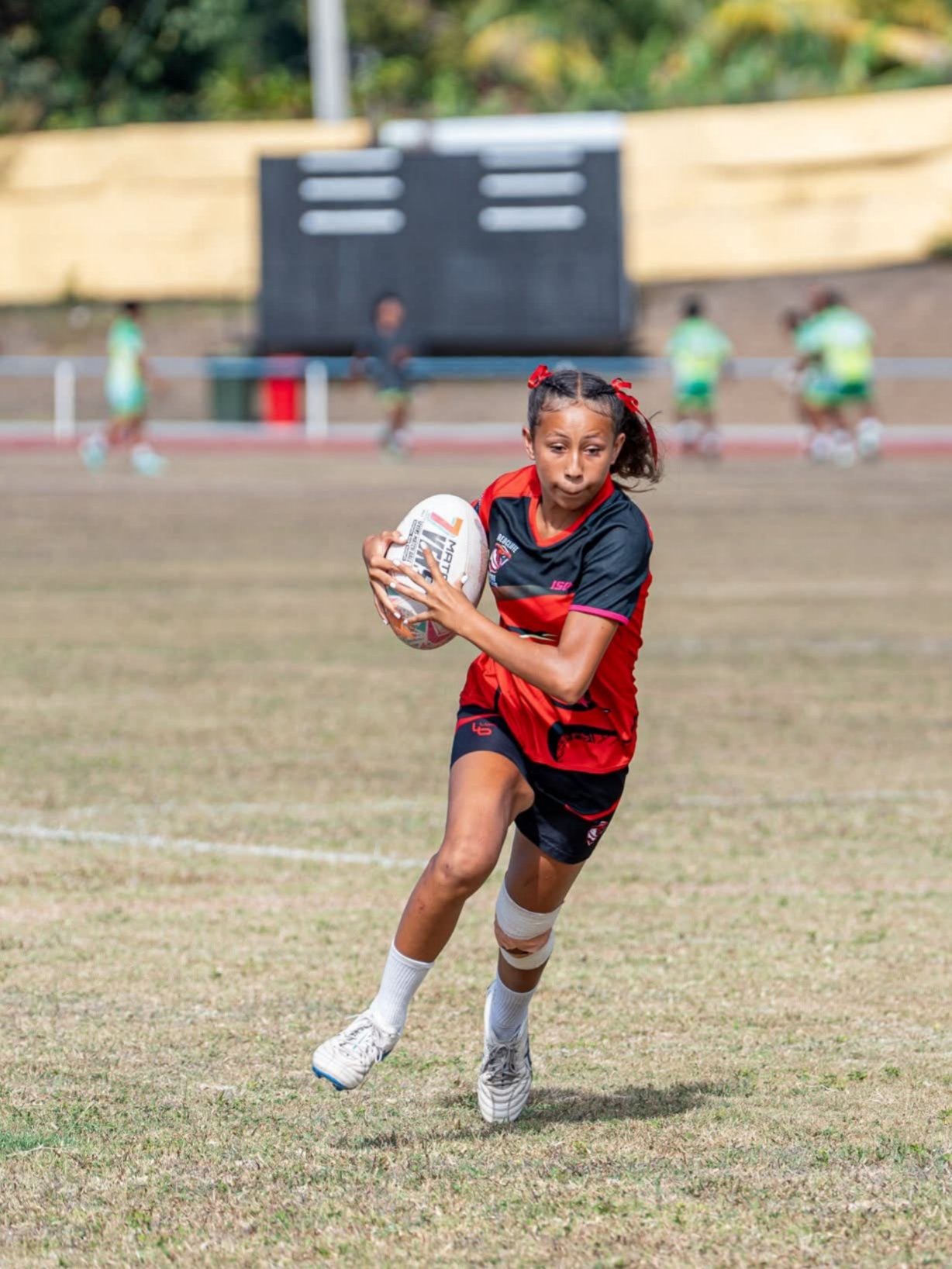 A young girl playing rugby on a field, holding a rugby ball, with other children in the background.