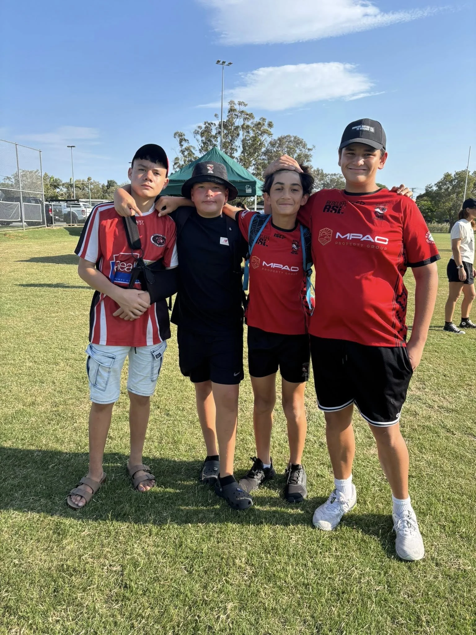 Four young boys standing on a grassy field, some wearing sports jerseys, smiling and posing for a photo with arm around each other during a sunny outdoor event.
