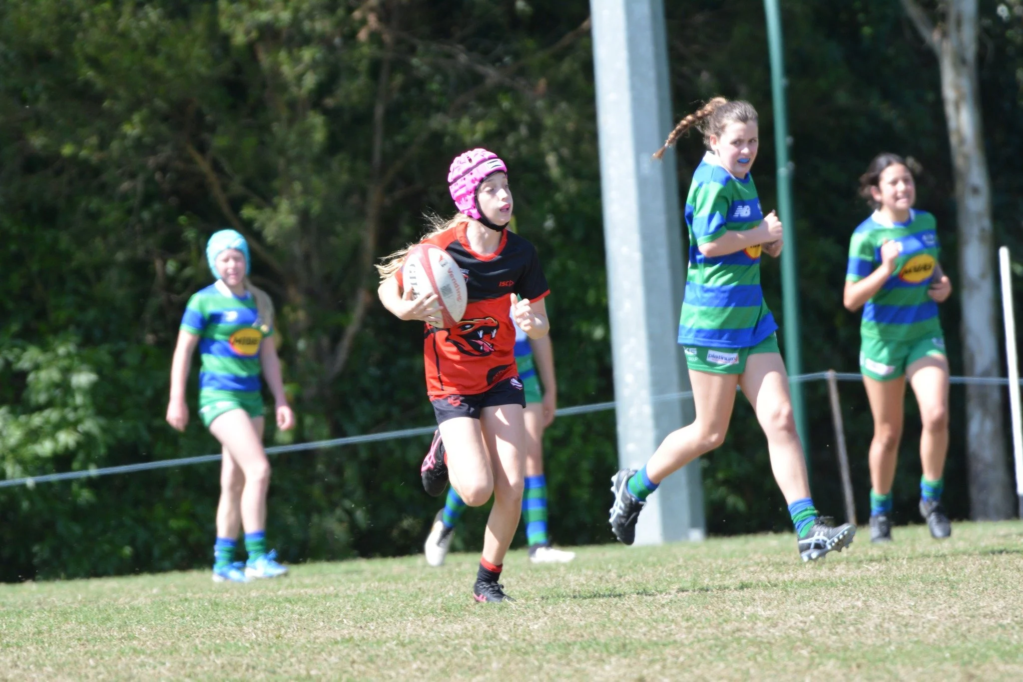 Girls playing rugby on a grassy field, one girl in red running with the ball and three girls in blue and green uniforms following behind.