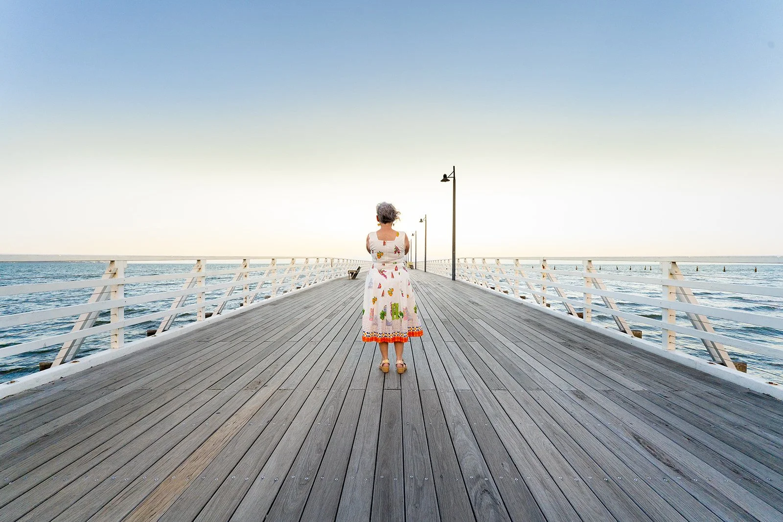 A woman in a white dress with colorful patterns stands on a wooden pier looking towards the horizon over the ocean during sunset or sunrise.