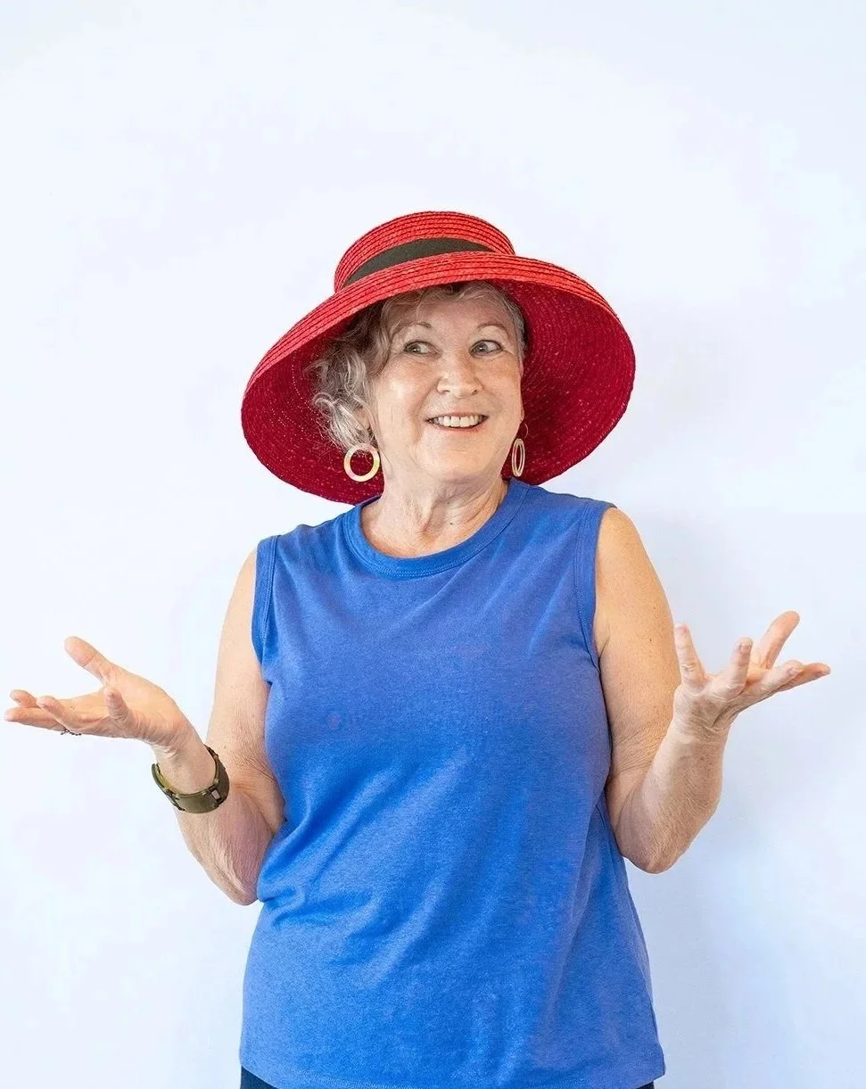 A woman in a blue sleeveless top wearing large earrings and a red wide-brimmed hat, smiling and gesturing with her hands against a plain white background.