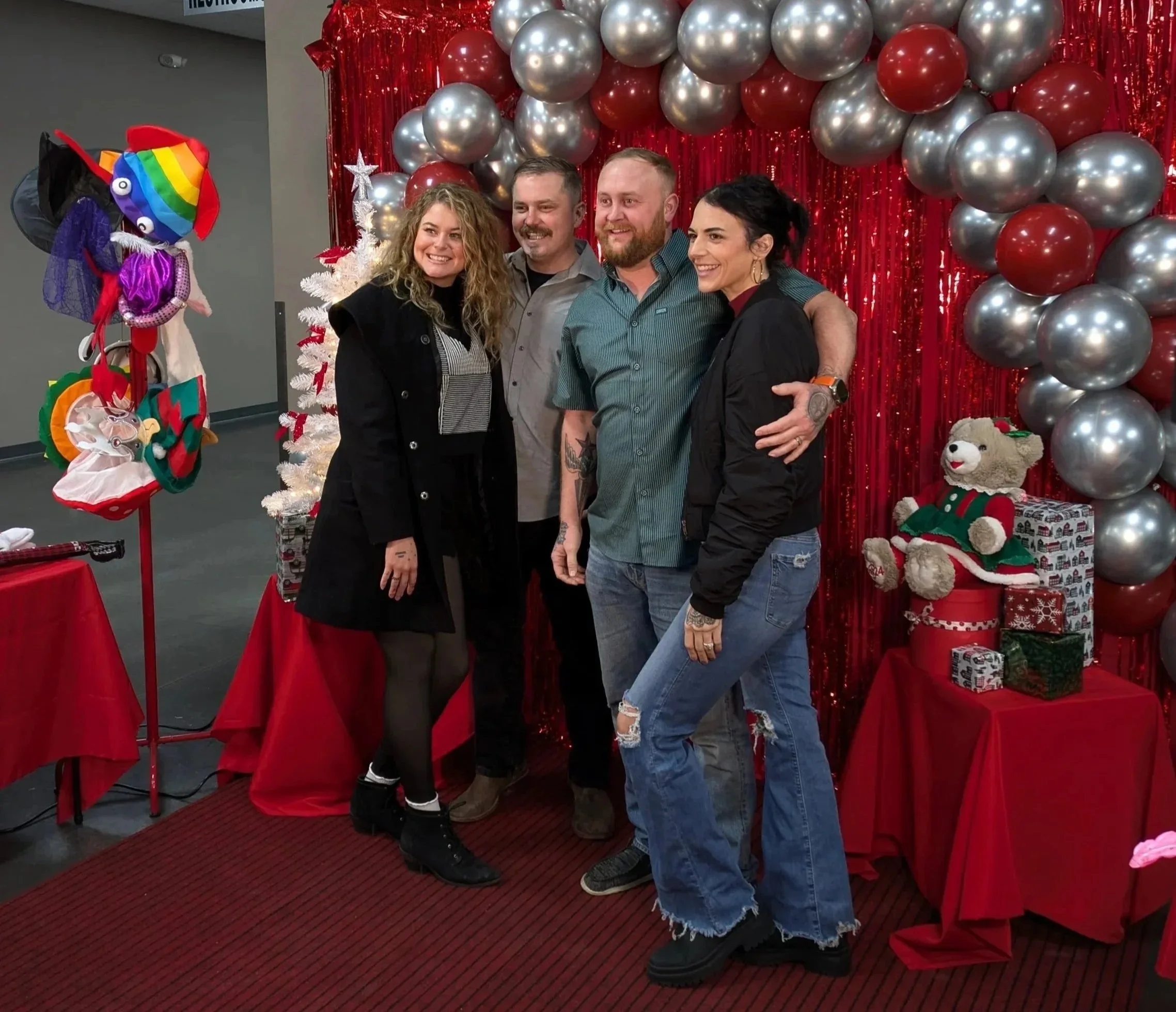 Four people standing together in front of a Christmas-themed backdrop with red and silver balloons, a white Christmas tree, and holiday decorations, smiling and posing for a photo.