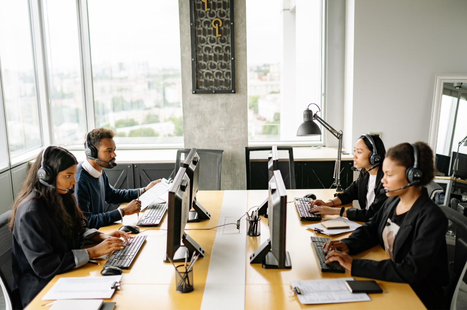 Four person team in a modern office with large windows, wearing headsets and working on desktop computers at a conference table.