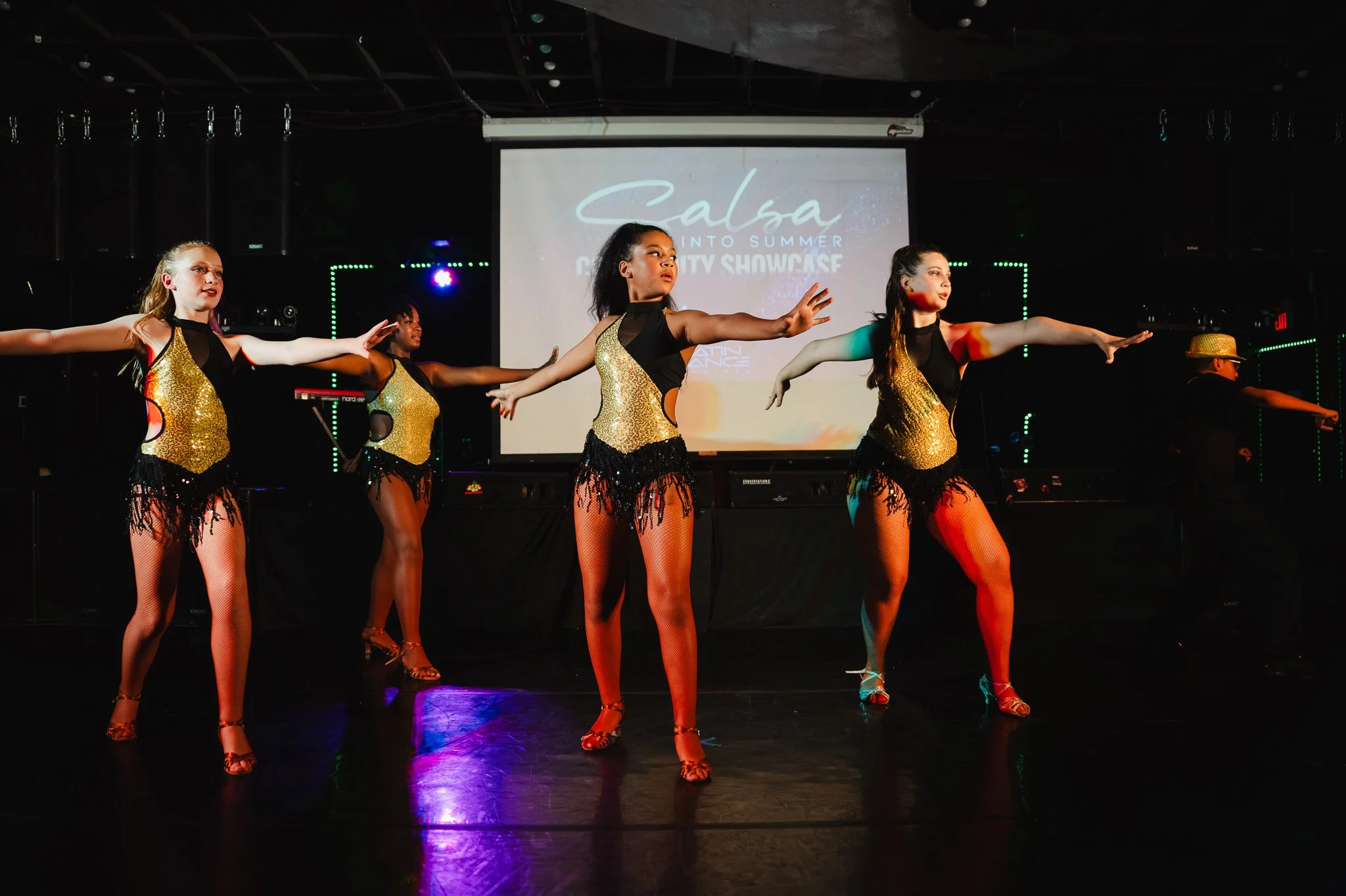 Children performing on stage in dark blue costumes with sequins, one boy with glasses and a hat, girls smiling and dancing.