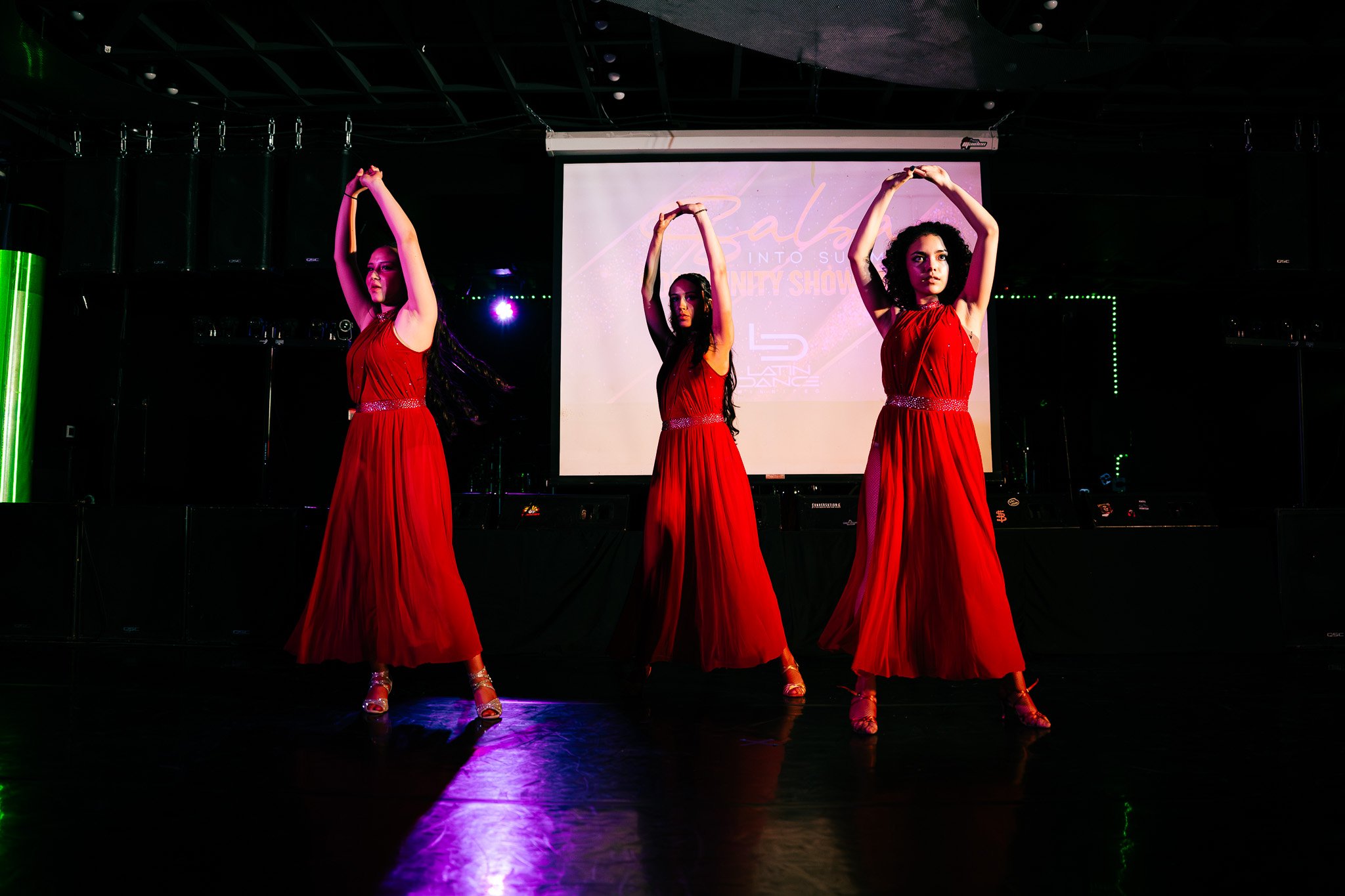 Three women dressed in black and leopard print costumes, performing a dance with headbands and hoop earrings, illuminated by stage lights.