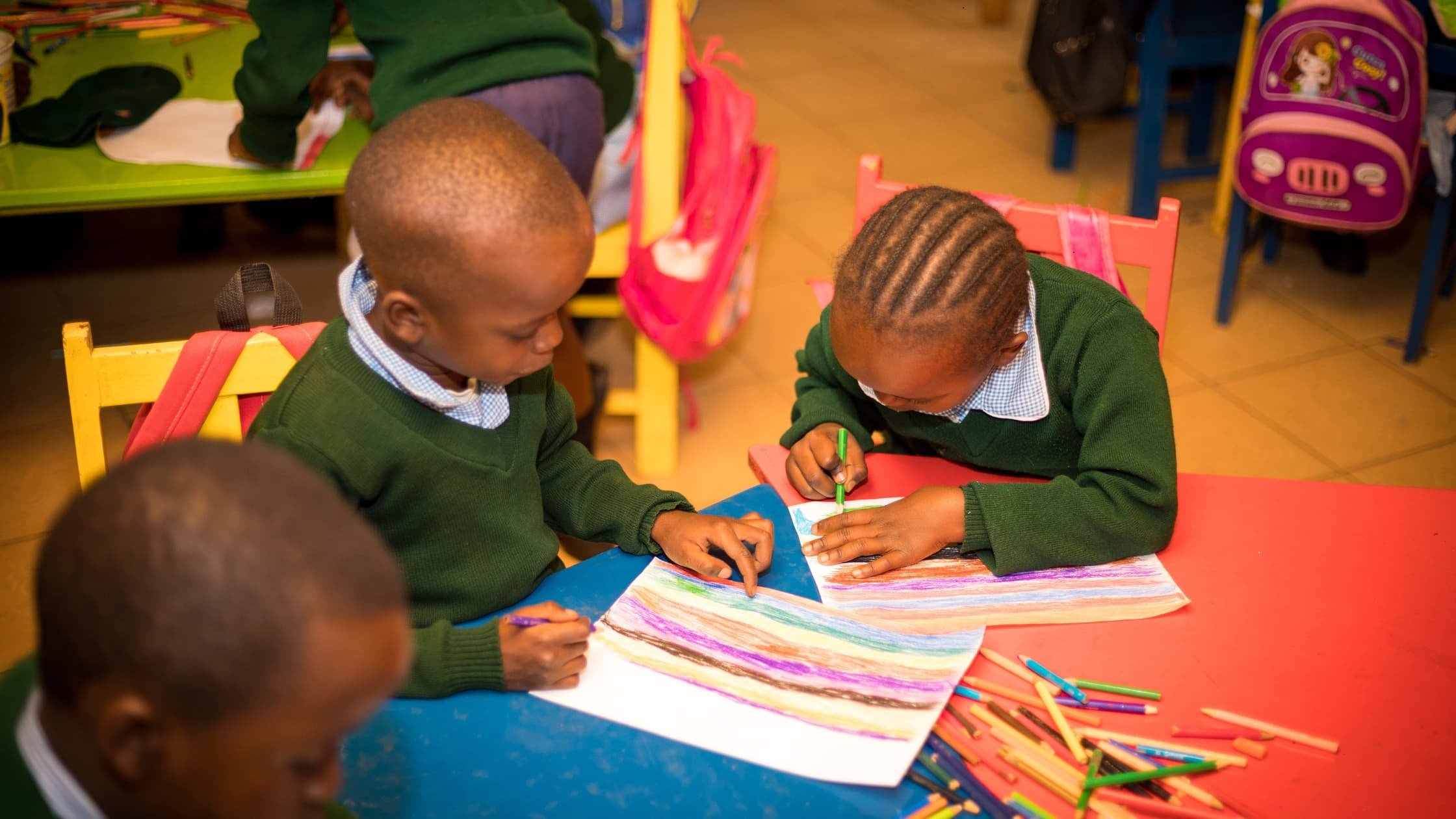 Three children in green school uniforms drawing and coloring on a large sheet of paper with colored pencils, with backpacks hanging on their chairs in a classroom.