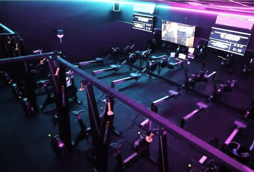 The Revelate room at The Engine Room Fitness, Ballincollig - Concept 2 rowers, ski ergs and bikes under purple and cyan lighting, with the class screens lit.