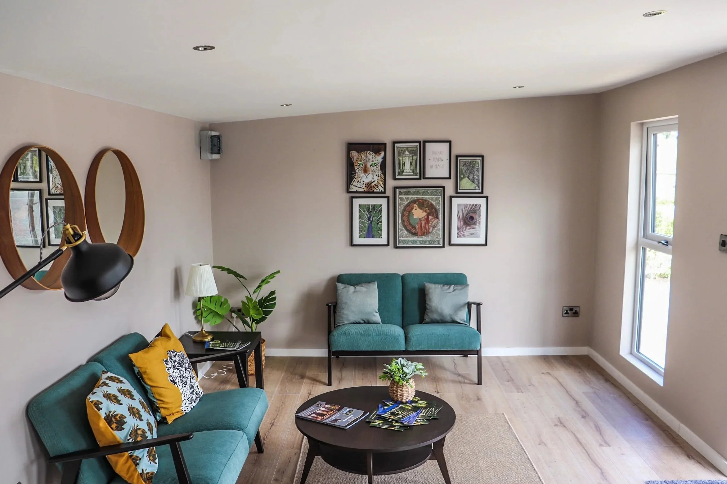 Living room with teal sofas, framed art on the wall, a black round coffee table with magazines and a potted plant, a side table with a white lamp, a plant, and decor accents, natural light from windows, and wall mirrors.