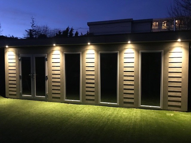 Exterior of a modern building with five glass doors, illuminated by outdoor lights at night, and a grass yard in front.