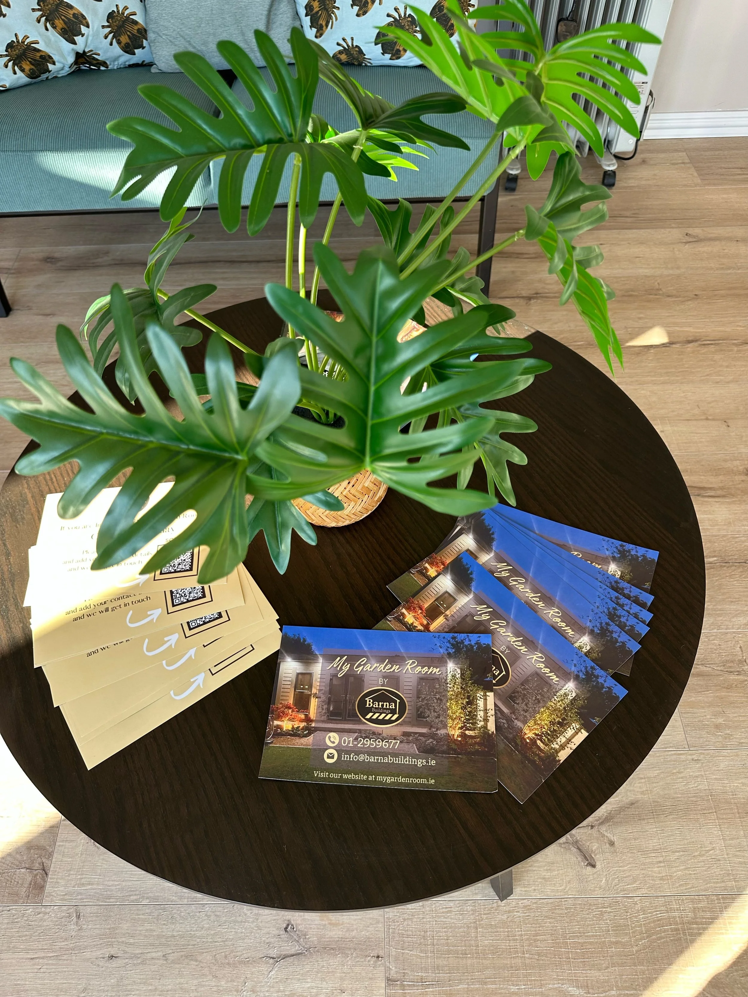 A round dark wooden table with a large potted monstera plant and business flyers for 'My Garden Room' from Barnal Building, featuring contact information and website, in a room with wooden flooring and a light blue sofa with patterned pillows.