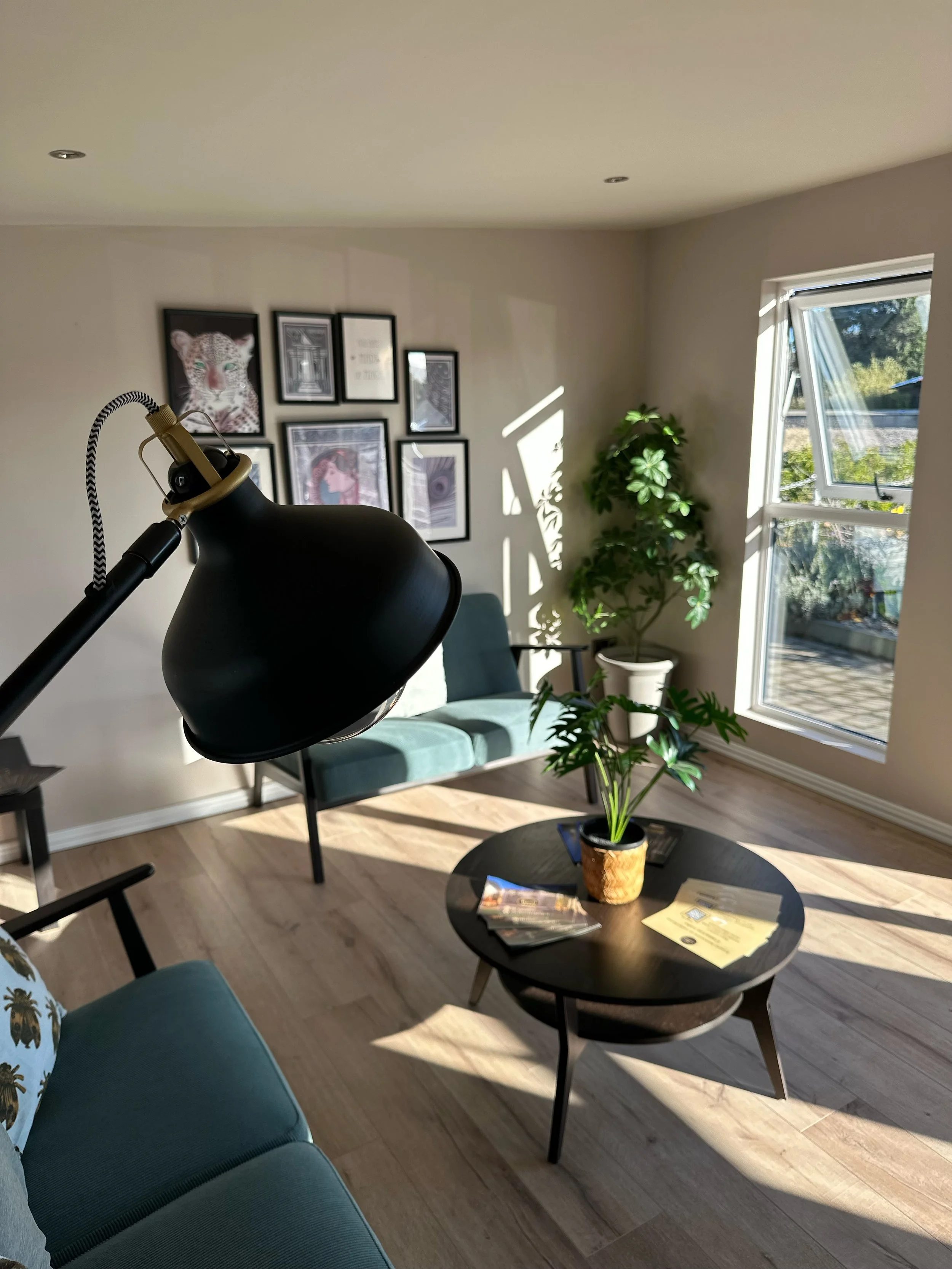 Living room with a round black coffee table, a potted plant, and a group of pictures on the wall. Sunlight streams through a window, creating shadows on the light-colored wooden floor.