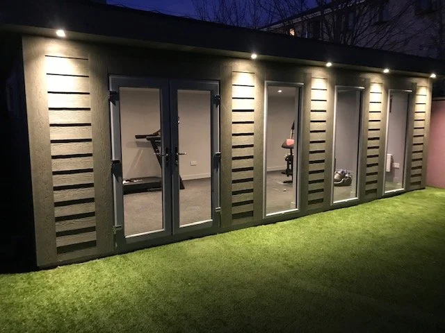 A modern backyard gym shed with glass doors and interior workout equipment at night.