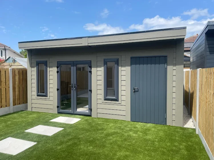 A small, modern backyard shed with gray siding, black window frames, a glass double door, and a separate storage door, surrounded by a well-maintained lawn and a wooden fence.