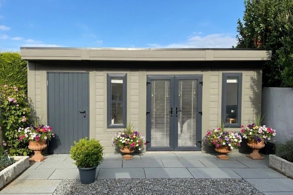 A small modern backyard shed painted in gray with double French doors and narrow side windows, surrounded by colorful potted flowers and greenery under a blue sky.