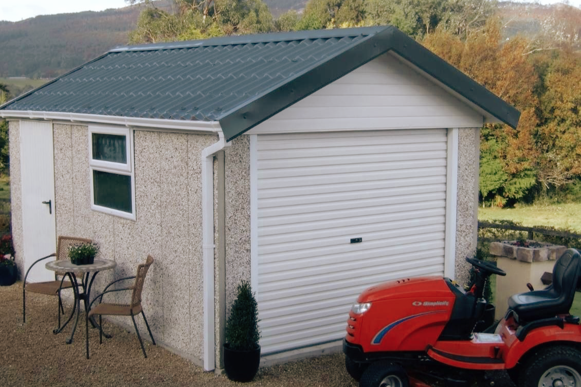 A small detached garage with a rolling shutter door, two windows, and a sitting area with two chairs and a small round table outside. A red lawn tractor is parked nearby, and the background features trees and hills.