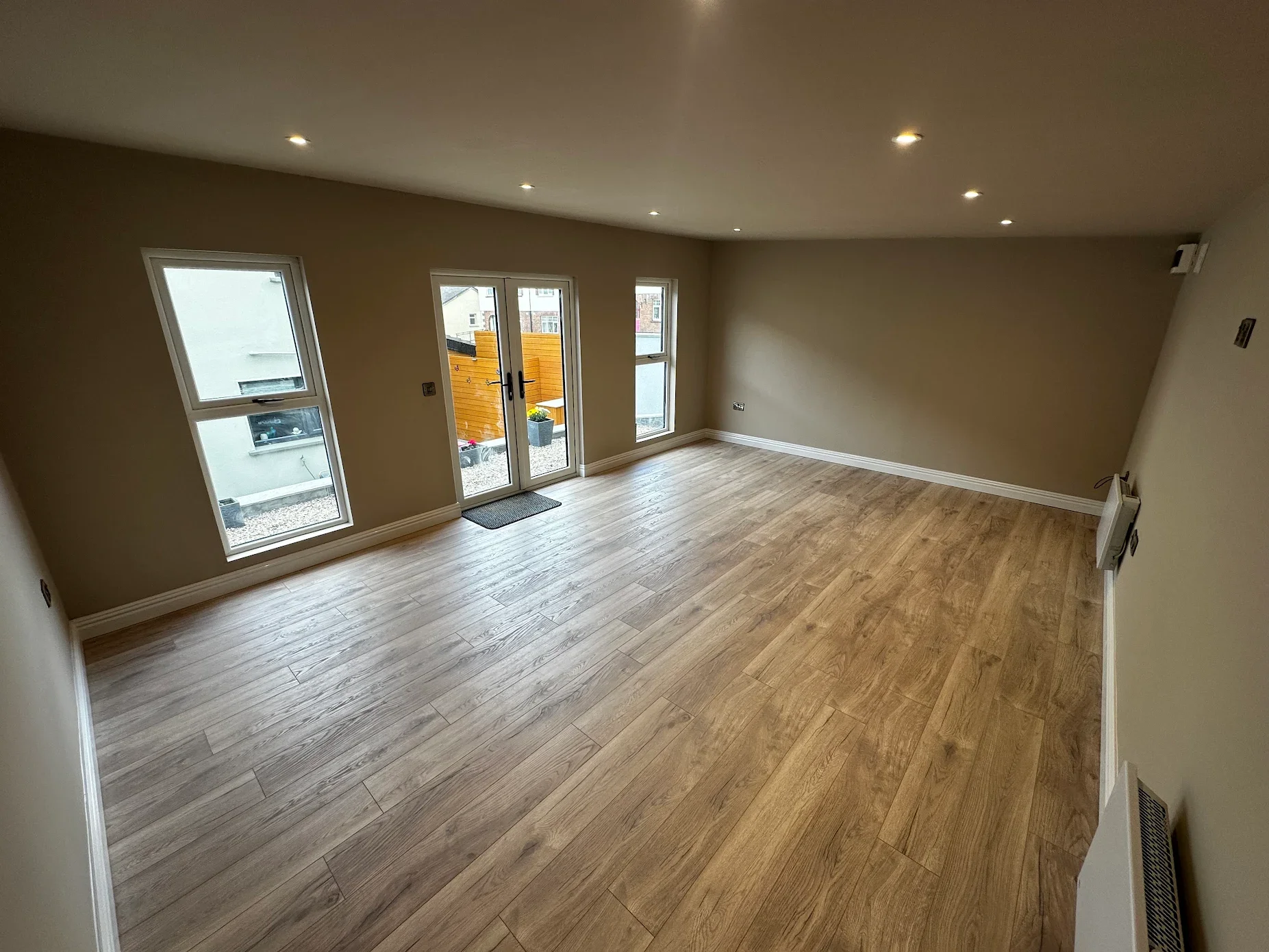 Empty living room with wooden flooring, beige walls, and three large windows with sliding glass door leading outside.