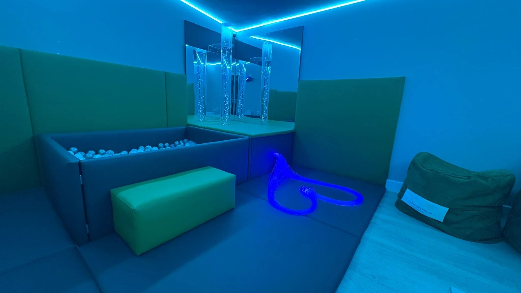 Indoor play area with neon blue lighting, padded green and blue walls, plastic ball pit, neon blue rope, and a bean bag chair on the wooden floor.