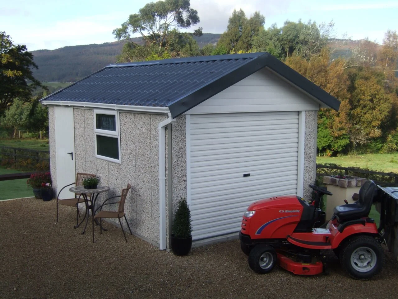 Small backyard shed with a black metal roof, white trim, a window, a white garage door, and outdoor furniture with a lawnmower parked outside, surrounded by trees and mountains.