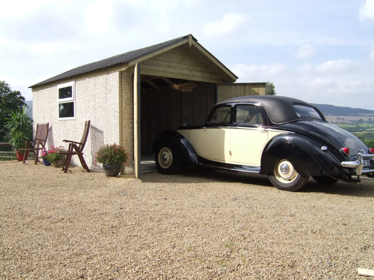Vintage black and white car parked in front of a wooden shed with potted plants and chairs outside on gravel, with rolling hills in the background.