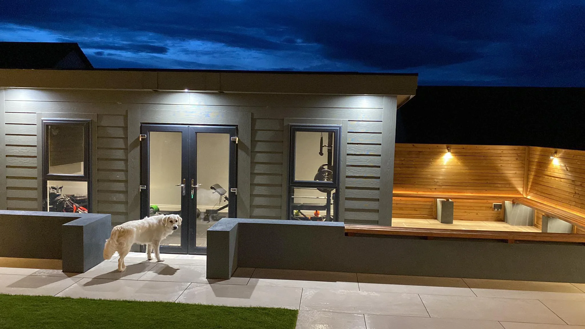A white dog standing on a patio outside a modern house at night. The house has large glass doors and windows, with outdoor lighting illuminating the area. There is a fenced-in wooden area with built-in benches to the right.