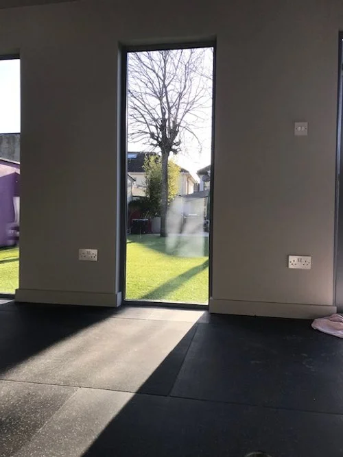 Interior view of a room with a glass door showing an outdoor yard and trees, sunlight casting shadows inside, with electrical outlets on the wall.