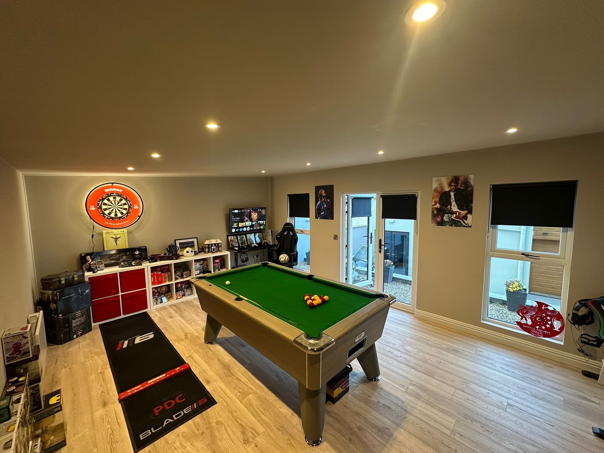 A game room with a pool table at the center, featuring billiard balls arranged in a triangle. The room has wooden flooring, black window blinds, and wall art. In the corner, there is a dartboard, a gaming chair, shelves with collectibles, and a TV. T