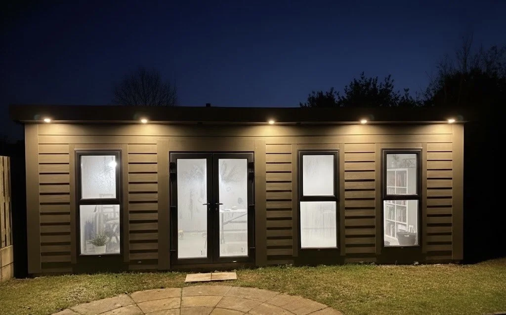 A small backyard shed with exterior lights on at dusk. The shed has brown horizontal siding, a pair of glass doors, and four windows, one on each side of the doors, with frosted or semi-transparent glass.