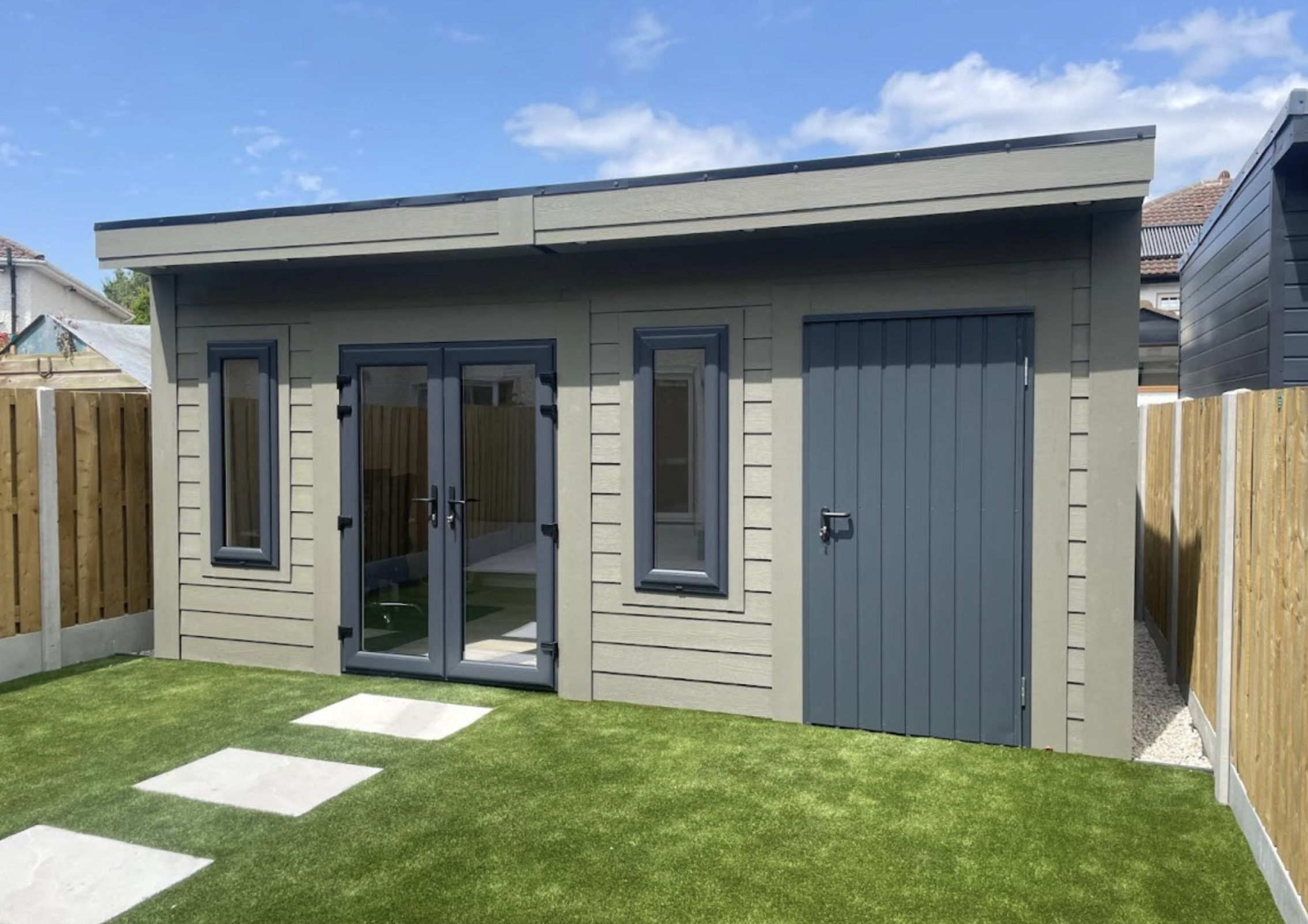 Grey garden shed with blue doors and windows, situated in a backyard with artificial grass and wooden fence.