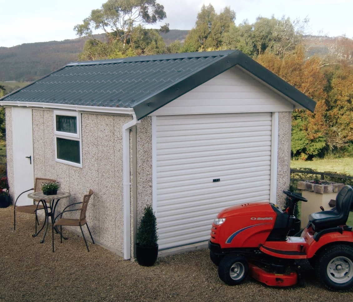 Small detached garage with white roll-up door, window, and seating area with two chairs and a small table outside. A red riding lawn mower is parked in front, and there are trees and hills in the background.