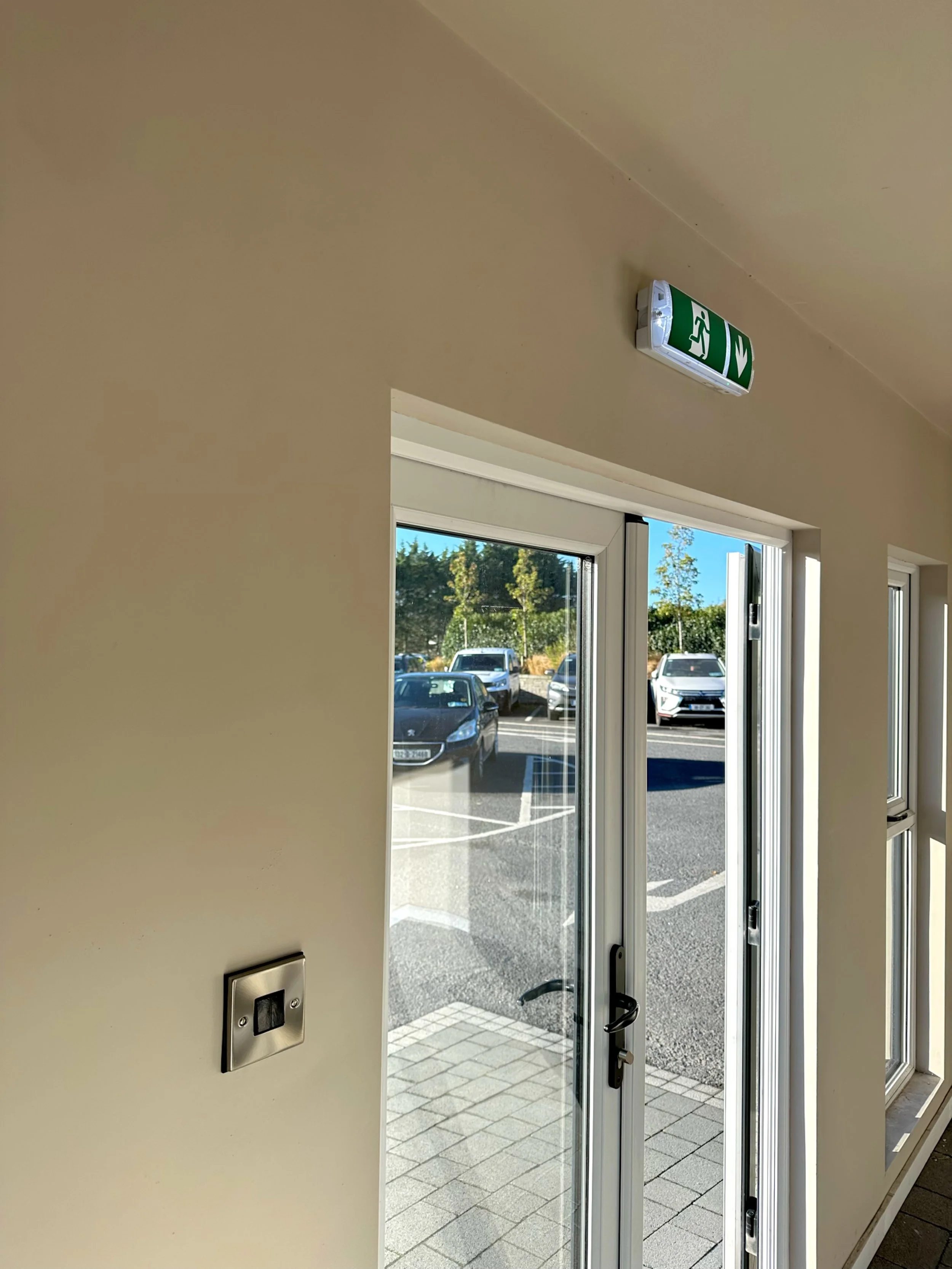 Interior view of a glass door leading outside to a parking lot with several cars, under a green and white emergency exit sign on a beige wall.