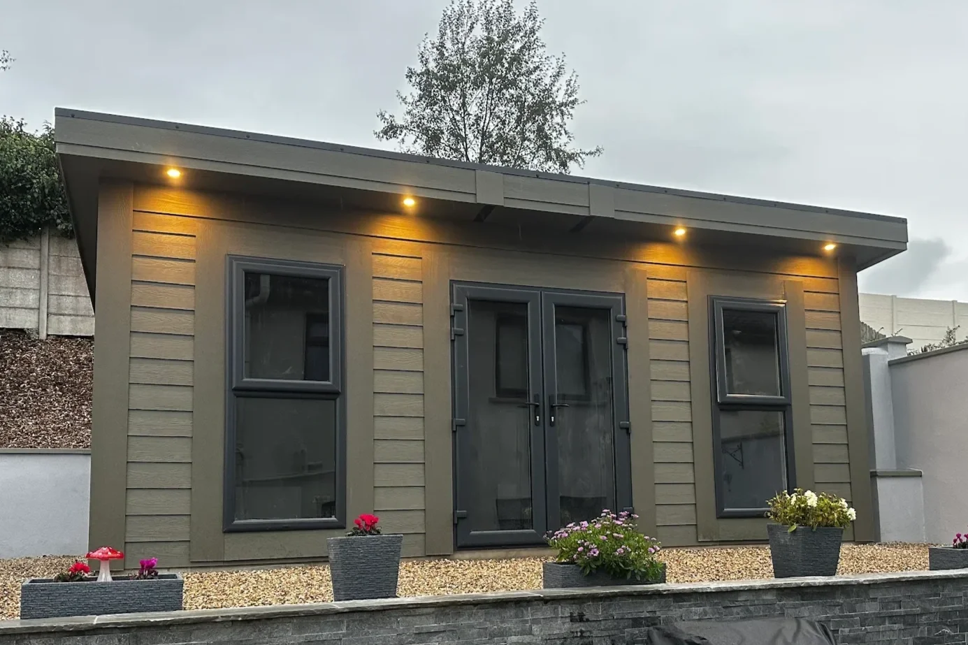 Small modern house with gray exterior, double glass door, two windows, and outdoor flower pots, at dusk with exterior lights on.