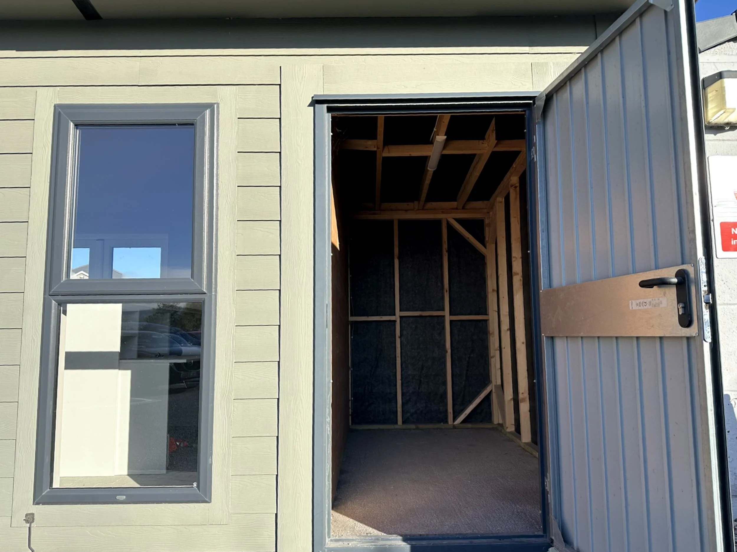 View of a building with a window on the left and an open door on the right, revealing an unfinished interior with wooden framing.
