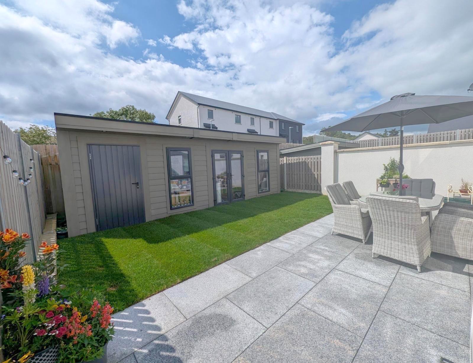 A backyard with a patio featuring a round table, eight wicker chairs, and a large umbrella. There is a small patch of lawn next to a shed with gray siding and black trim, and a flower bed with colorful flowers in the foreground. The backyard is enclo